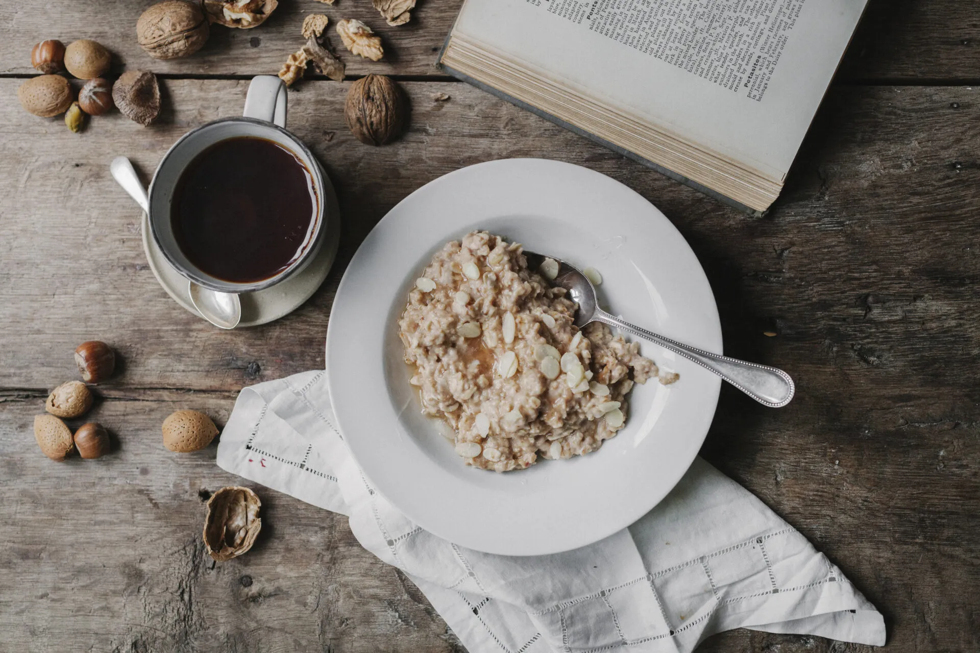 Piatto di porridge d'avena con mandorle a lamelle accanto a una tazza di caffè nero, frutta secca e tovagliolo di lino su un tavolo di legno