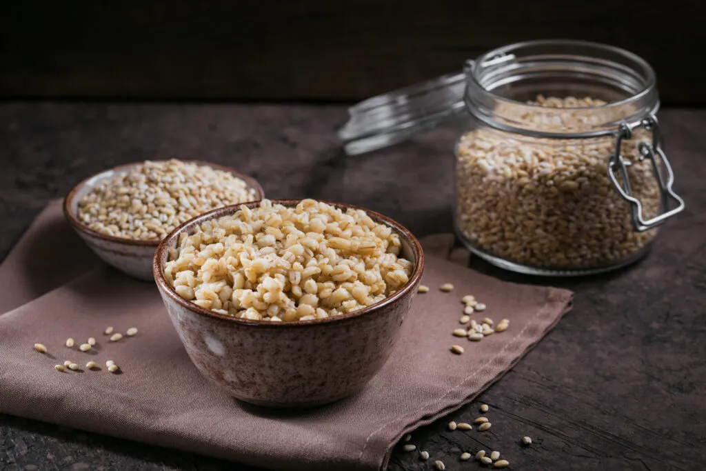 Ciotola di orzo perlato cotto in primo piano, con una ciotolina di chicchi crudi e un barattolo di vetro sullo sfondo