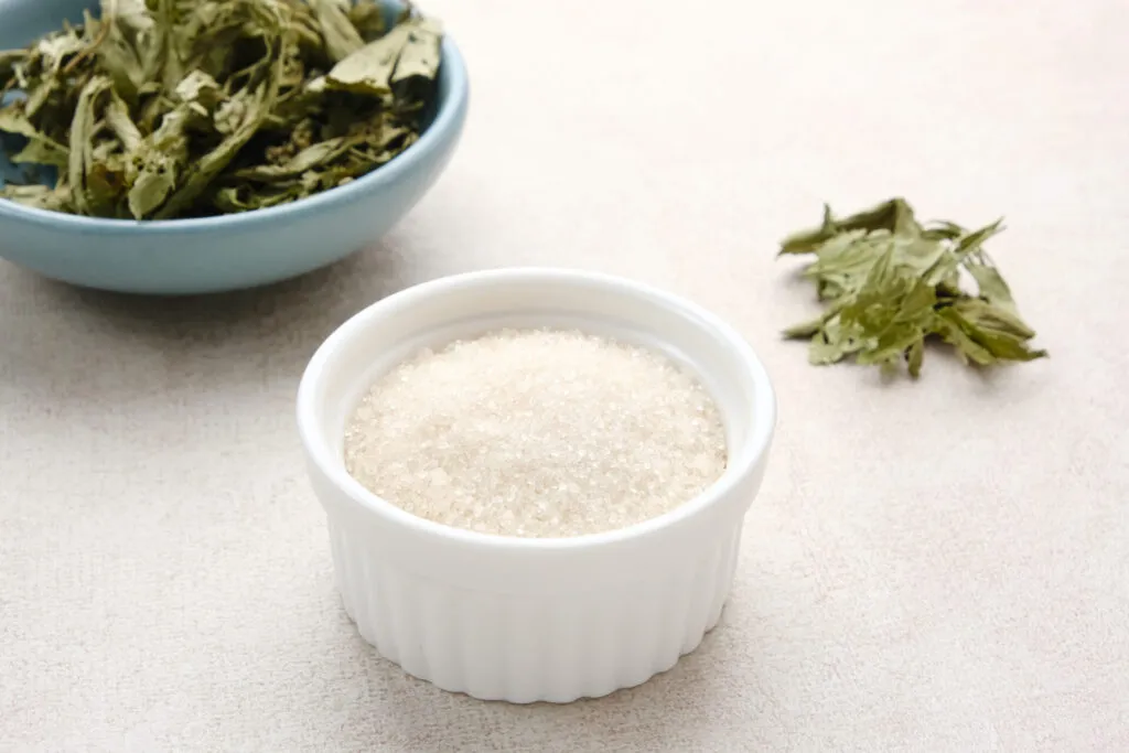 Ciotolina bianca con dolcificante alla stevia in cristalli, accanto a una ciotola azzurra di foglie di stevia essiccate su fondo chiaro