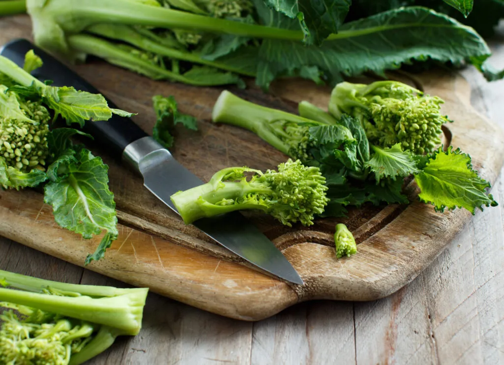 Cime di rapa in fase di pulizia su un tagliere di legno, con foglie tenere, cimette e gambi separati accanto a un coltello