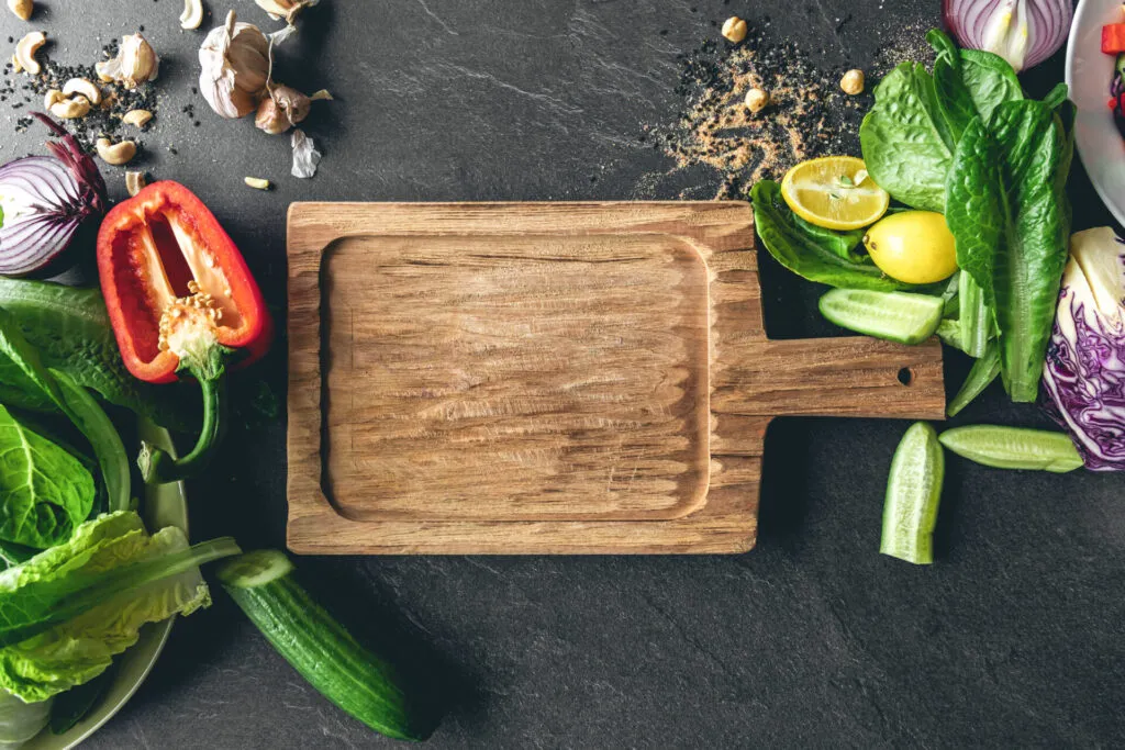 Tagliere di legno vuoto visto dall'alto, circondato da verdure fresche, limoni, aglio e frutta secca su fondo di ardesia scura