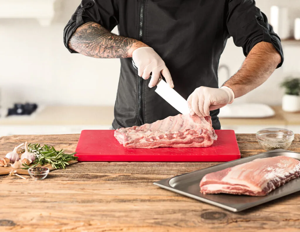 Cuoco con guanti che taglia carne di maiale cruda su un tagliere di plastica rosso, appoggiato su un piano di legno