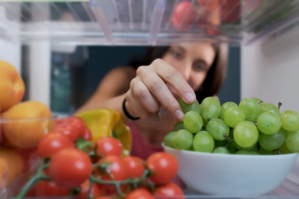 Mano che prende un acino da un grappolo d'uva bianca in una ciotola aperta dentro il frigorifero