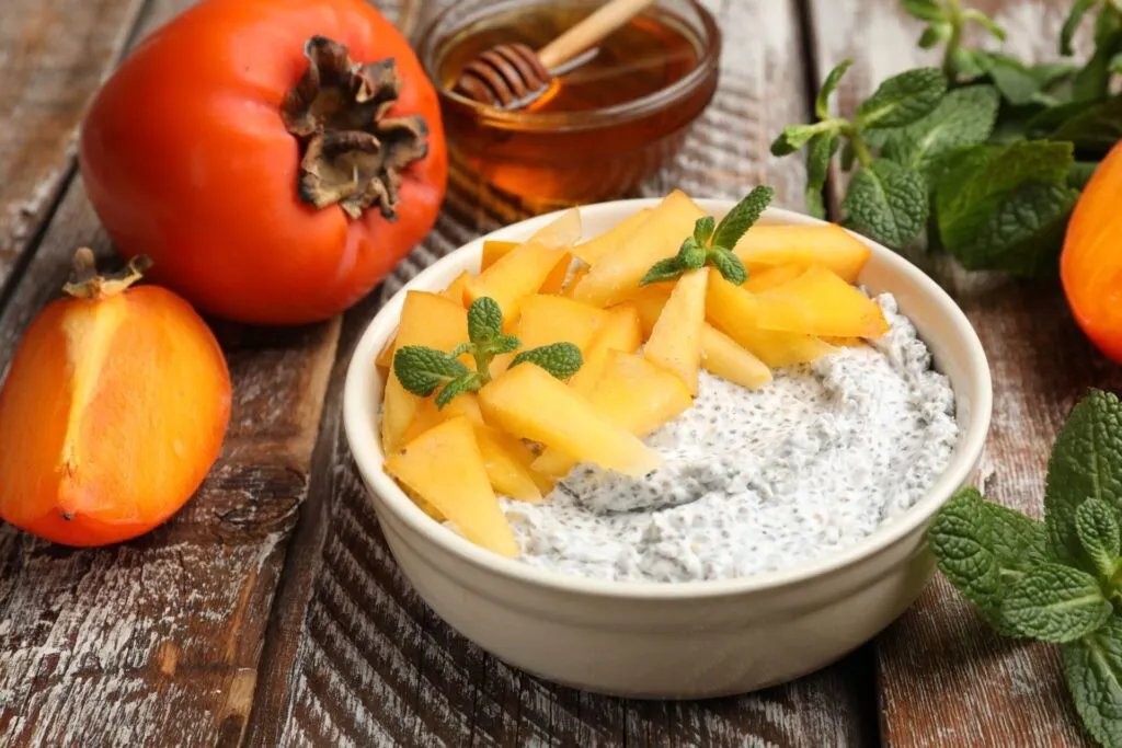 Ciotola di crema bianca con semi di chia guarnita con spicchi di cachi e foglioline di menta, accanto a un cachi intero e a uno tagliato a metà