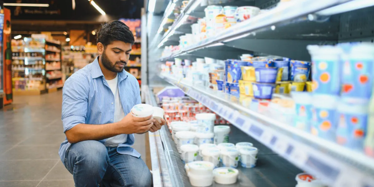 Persona accovacciata davanti al banco frigo del supermercato che tiene due vasetti bianchi, uno per mano, confrontandoli prima di scegliere