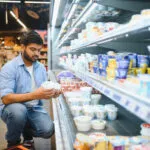 Persona accovacciata davanti al banco frigo del supermercato che tiene due vasetti bianchi, uno per mano, confrontandoli prima di scegliere