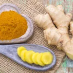 Rizoma di zenzero fresco accanto a una ciotola di legno con curcuma in polvere e fettine di curcuma su un cucchiaio