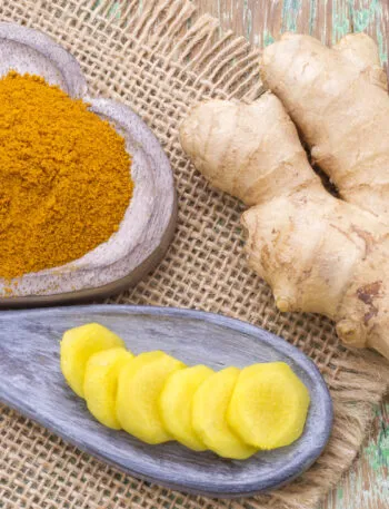 Rizoma di zenzero fresco accanto a una ciotola di legno con curcuma in polvere e fettine di curcuma su un cucchiaio