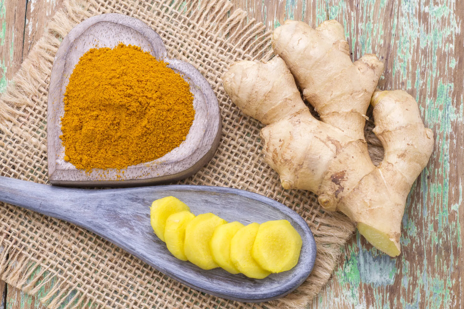 Rizoma di zenzero fresco accanto a una ciotola di legno con curcuma in polvere e fettine di curcuma su un cucchiaio