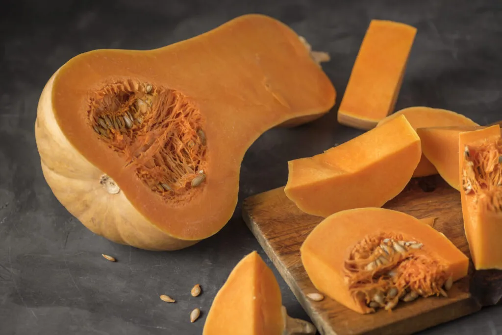 Zucca Butternut tagliata per il lungo, con i semi concentrati nella parte larga e il collo di sola polpa, accanto a fette su un tagliere