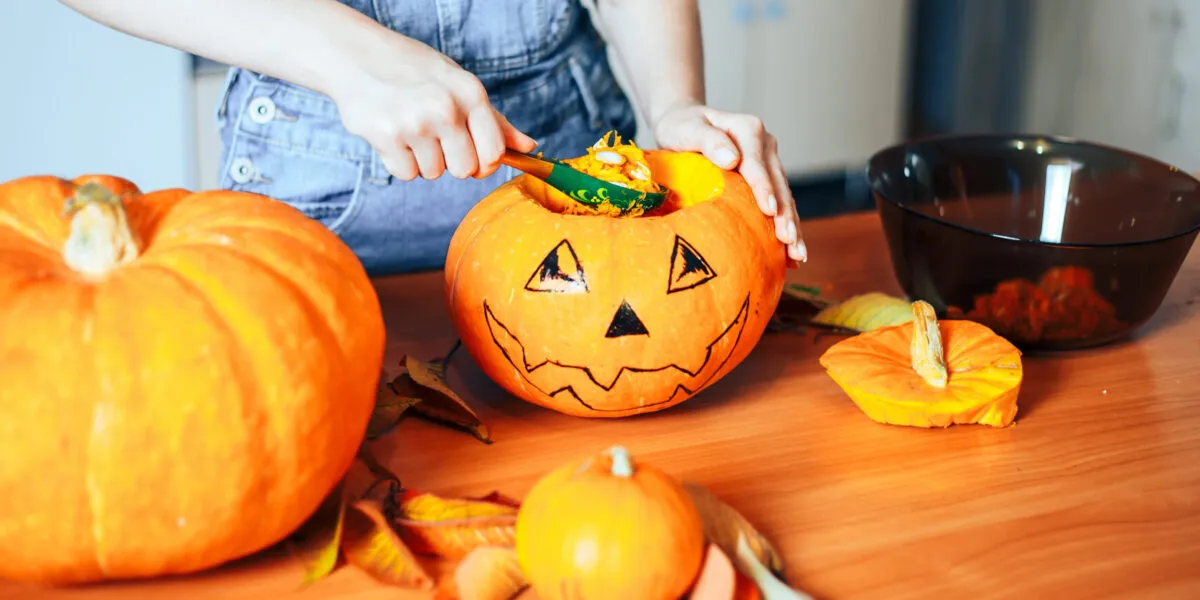Mani che svuotano con un cucchiaio una zucca di Halloween decorata, con la polpa arancione raccolta in una ciotola accanto e altre zucche sul tavolo