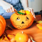 Mani che svuotano con un cucchiaio una zucca di Halloween decorata, con la polpa arancione raccolta in una ciotola accanto e altre zucche sul tavolo