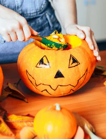 Mani che svuotano con un cucchiaio una zucca di Halloween decorata, con la polpa arancione raccolta in una ciotola accanto e altre zucche sul tavolo