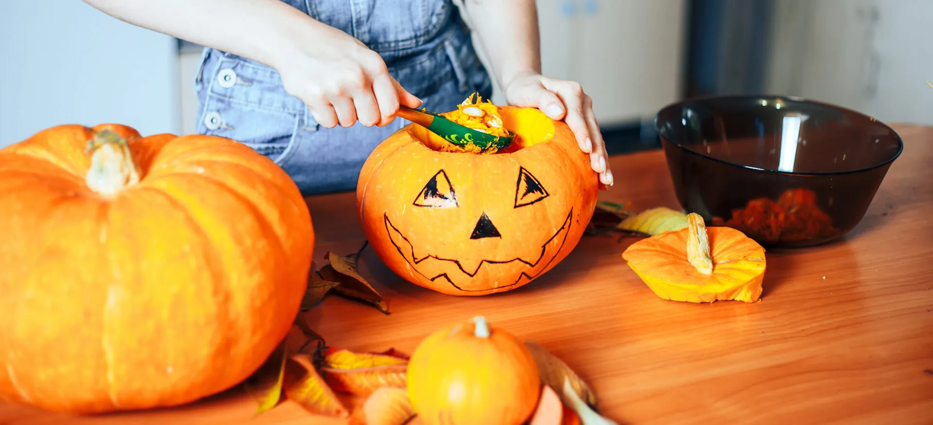 Mani che svuotano con un cucchiaio una zucca di Halloween decorata, con la polpa arancione raccolta in una ciotola accanto e altre zucche sul tavolo
