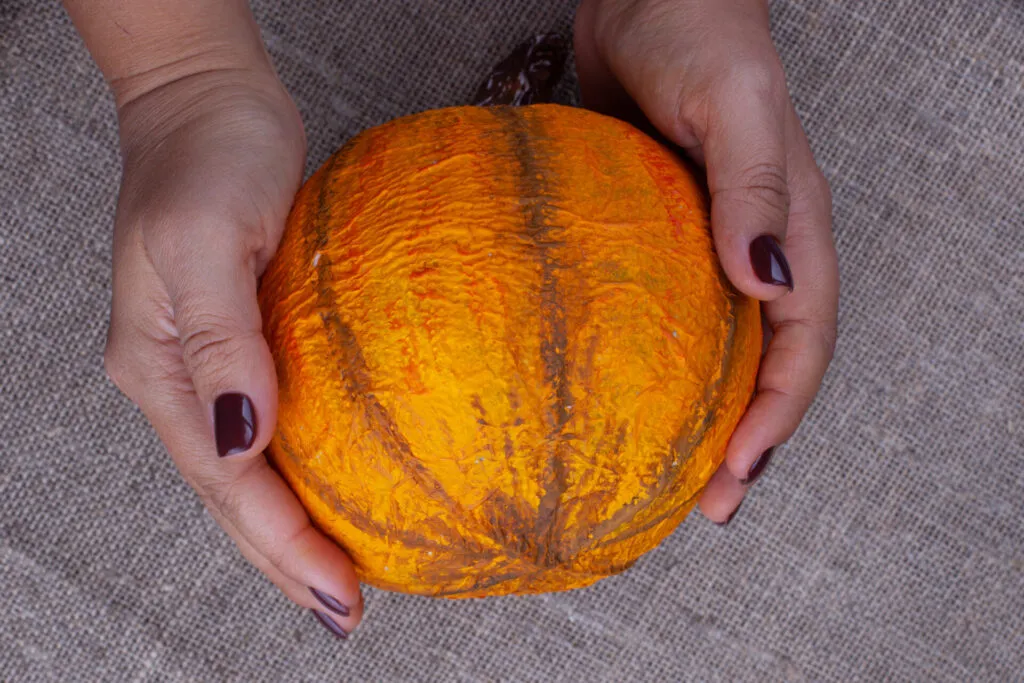 Due mani che tengono una piccola zucca arancione, con la scorza spessa e costolata ben visibile, su un fondo di juta