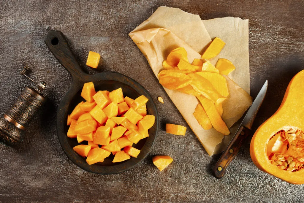 Cubetti di zucca arancione in un recipiente di legno scuro, con le bucce su carta forno e un coltello accanto
