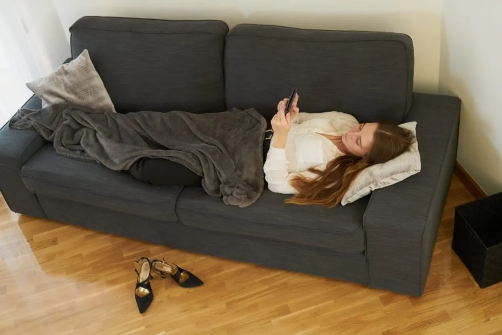 Donna sdraiata sul divano con una coperta e il telefono in mano, con le scarpe col tacco tolte sul pavimento