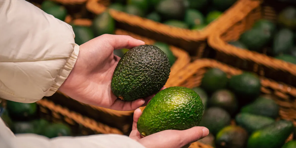 Due mani femminili reggono due avocado di colore diverso, uno verde scuro e uno verde chiaro, davanti al banco della frutta