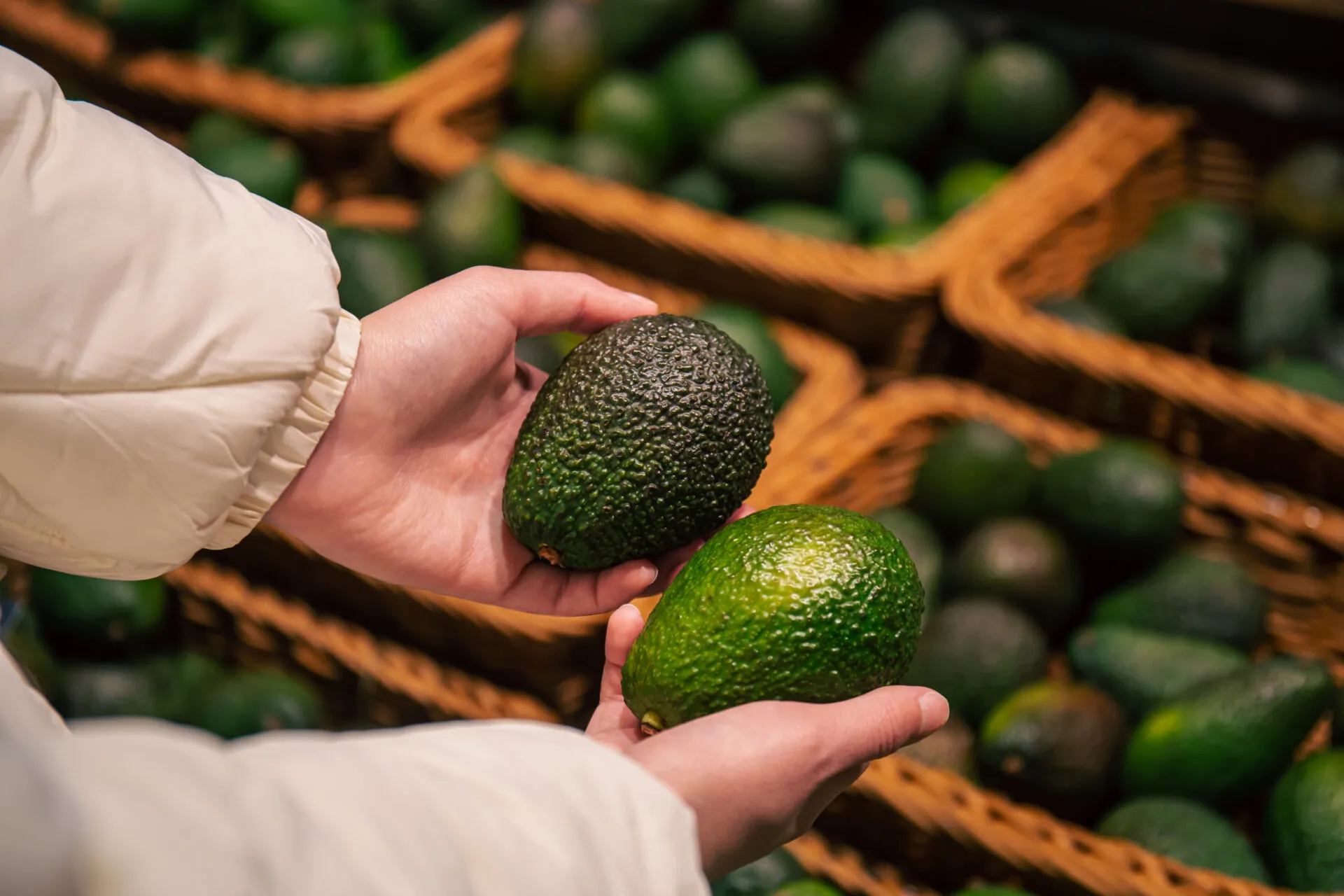 Due mani femminili reggono due avocado di colore diverso, uno verde scuro e uno verde chiaro, davanti al banco della frutta