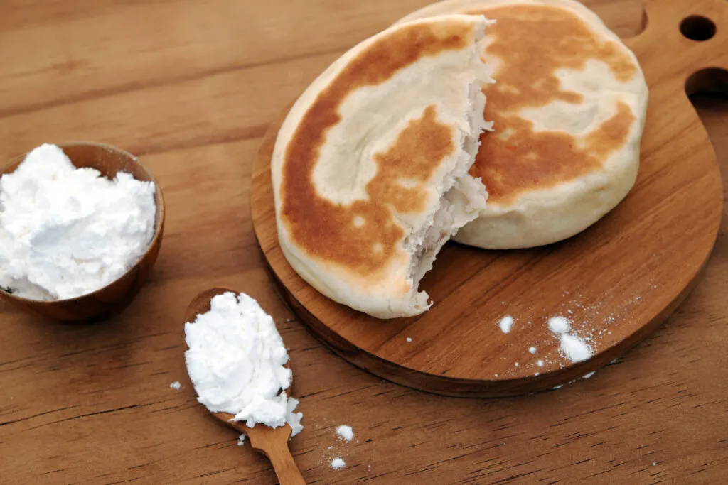 Focaccine cotte in padella su un tagliere di legno, una spezzata a metà con la mollica soffice, accanto a una ciotolina e un cucchiaio di polvere bianca
