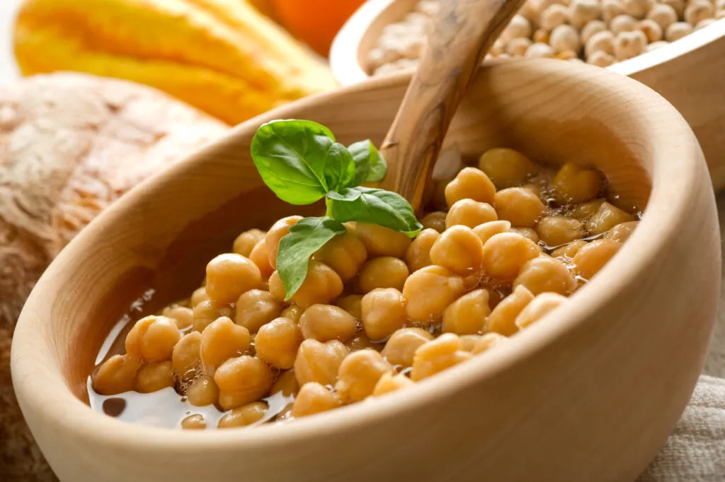 Ceci cotti nel loro liquido in una ciotola di legno con un cucchiaio di legno e foglie di basilico, ceci secchi sullo sfondo