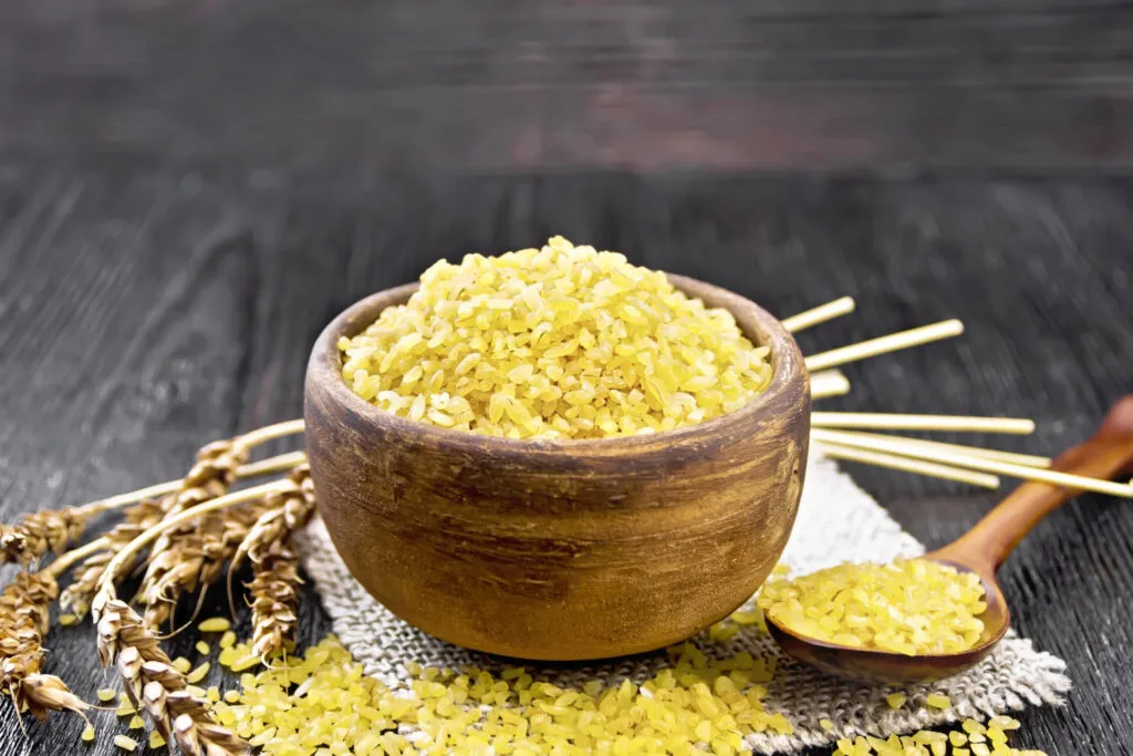 Bulgur crudo in una ciotola di legno con un cucchiaio e spighe di grano su un tavolo scuro