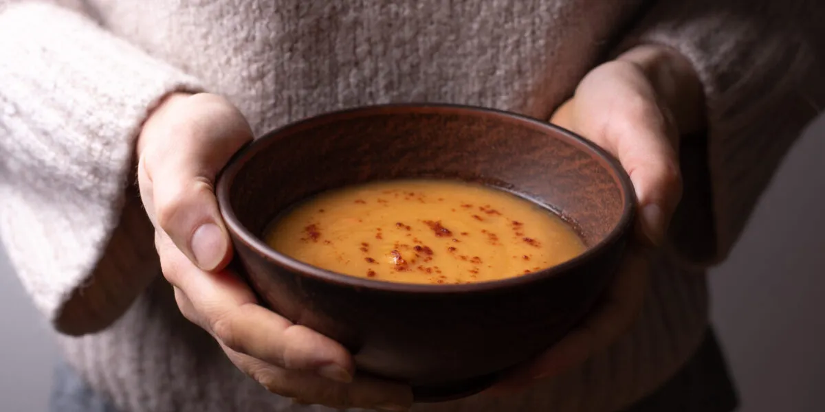 Mani che stringono una ciotola di ceramica scura con una vellutata arancione calda, spolverata di paprika