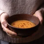Mani che stringono una ciotola di ceramica scura con una vellutata arancione calda, spolverata di paprika