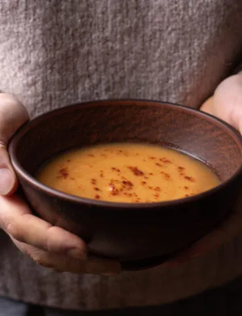 Mani che stringono una ciotola di ceramica scura con una vellutata arancione calda, spolverata di paprika