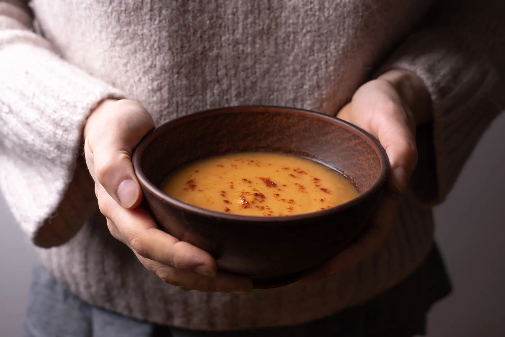 Mani che stringono una ciotola di ceramica scura con una vellutata arancione calda, spolverata di paprika