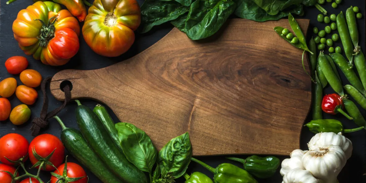 Verdure fresche intorno a un tagliere di legno: pomodori, peperoni, piselli, cetrioli, spinaci e aglio
