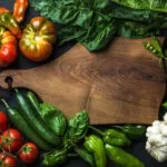 Verdure fresche intorno a un tagliere di legno: pomodori, peperoni, piselli, cetrioli, spinaci e aglio