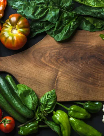 Verdure fresche intorno a un tagliere di legno: pomodori, peperoni, piselli, cetrioli, spinaci e aglio