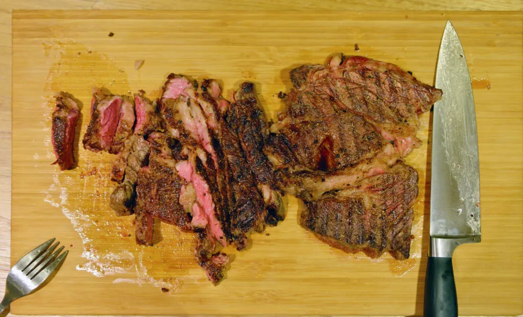 Bistecca cotta tagliata a fette su un tagliere di bambù, con liquido di cottura sparso intorno e un coltello da chef accanto.