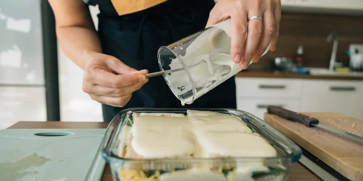 Mani femminili versano la besciamella da una brocca su una lasagna in una teglia di vetro, con gli strati di pasta e verdure visibili attraverso la parete