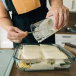 Mani femminili versano la besciamella da una brocca su una lasagna in una teglia di vetro, con gli strati di pasta e verdure visibili attraverso la parete