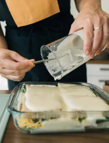 Mani femminili versano la besciamella da una brocca su una lasagna in una teglia di vetro, con gli strati di pasta e verdure visibili attraverso la parete