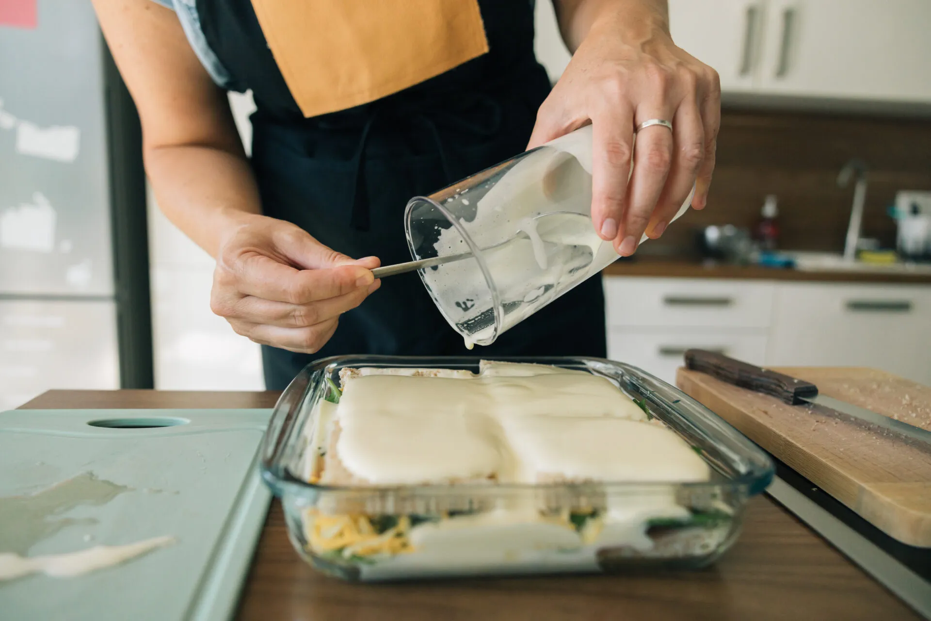 Mani femminili versano la besciamella da una brocca su una lasagna in una teglia di vetro, con gli strati di pasta e verdure visibili attraverso la parete