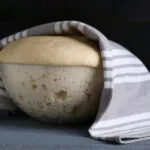 Impasto lievitato in una ciotola di vetro coperta da un canovaccio, con le bolle della lievitazione visibili attraverso la parete