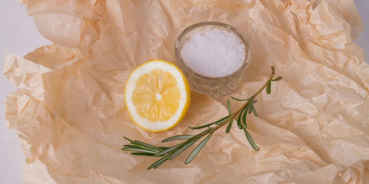 Mezzo limone e un rametto di rosmarino accanto a una ciotolina di sale grosso su carta forno