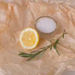 Mezzo limone e un rametto di rosmarino accanto a una ciotolina di sale grosso su carta forno