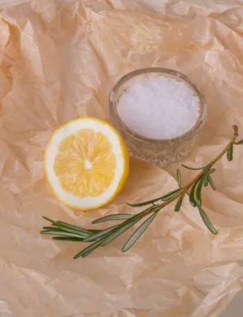 Mezzo limone e un rametto di rosmarino accanto a una ciotolina di sale grosso su carta forno