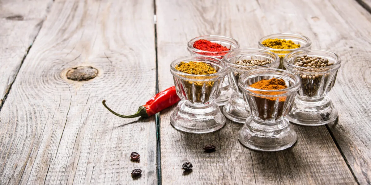 Spezie in polvere e in semi di colori diversi in sei bicchierini di vetro su un tavolo di legno, con un peperoncino rosso secco accanto