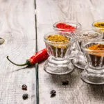 Spezie in polvere e in semi di colori diversi in sei bicchierini di vetro su un tavolo di legno, con un peperoncino rosso secco accanto